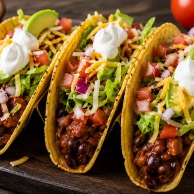 Three loaded chili tacos topped with lettuce, tomatoes, avocado, shredded cheese, red onion, and sour cream on a wooden serving tray.
