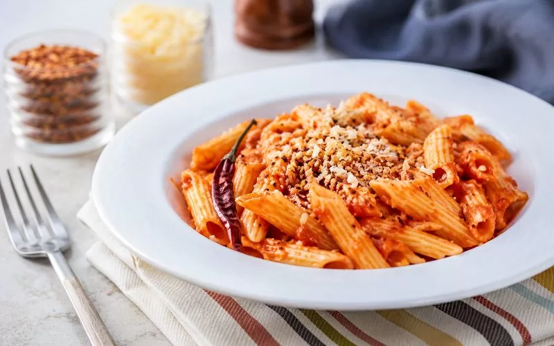 Spicy marinara penne pasta served in a white bowl with Parmesan cheese, red pepper flakes, and a dried chili pepper.