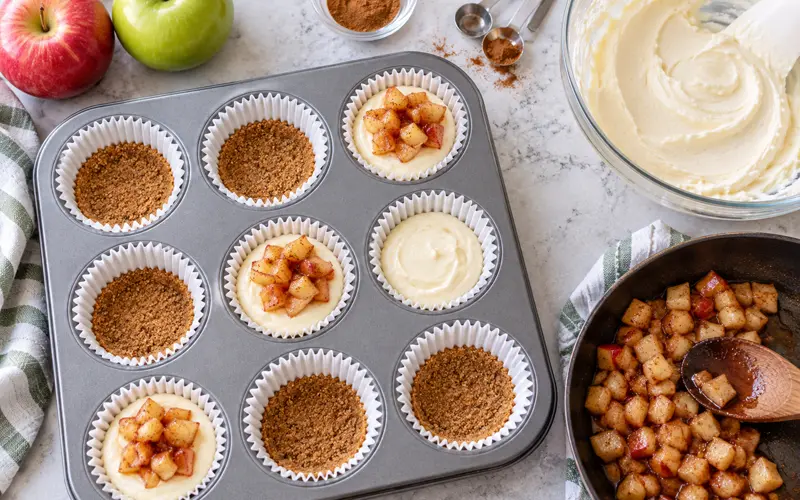 Preparing mini apple cheesecakes in a muffin pan with graham cracker crust, cheesecake filling, and cinnamon apples