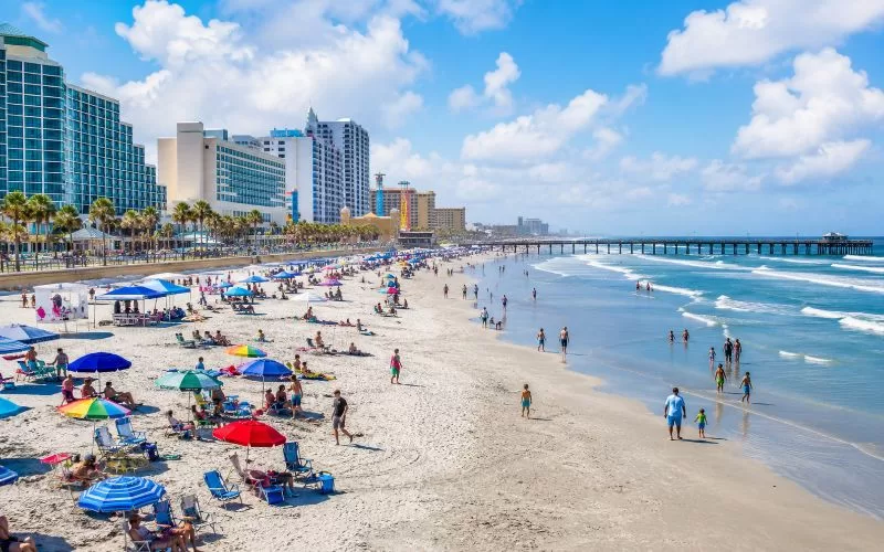 Busy Daytona Beach shoreline with colorful umbrellas, families, beachfront hotels, and a pier under a bright blue sky.