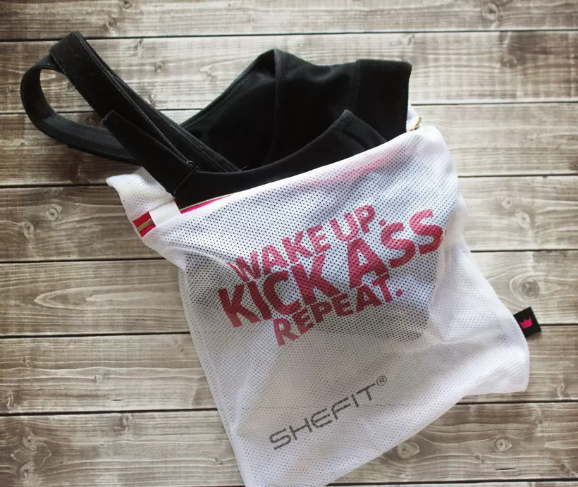 Black SHEFIT Ultimate Sports Bra partially placed inside a white mesh laundry bag.