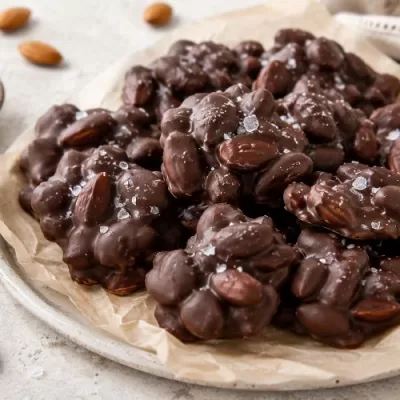 Dark chocolate almond clusters sprinkled with flaky sea salt on parchment with whole almonds and a small bowl of salt nearby.