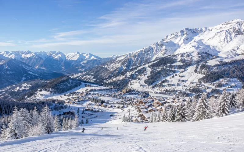 Snow-covered mountain ski resort with alpine slopes, a village below, and skiers in the distance.