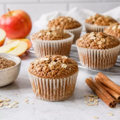 Freshly baked apple cinnamon flaxseed muffins with apples, oats, cinnamon sticks, and ground flaxseed on a light kitchen counter.