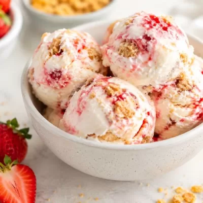 Bowl of homemade strawberry cheesecake ice cream with strawberry swirls, graham cracker pieces, fresh strawberries, and graham cracker crumbs on a light counter.