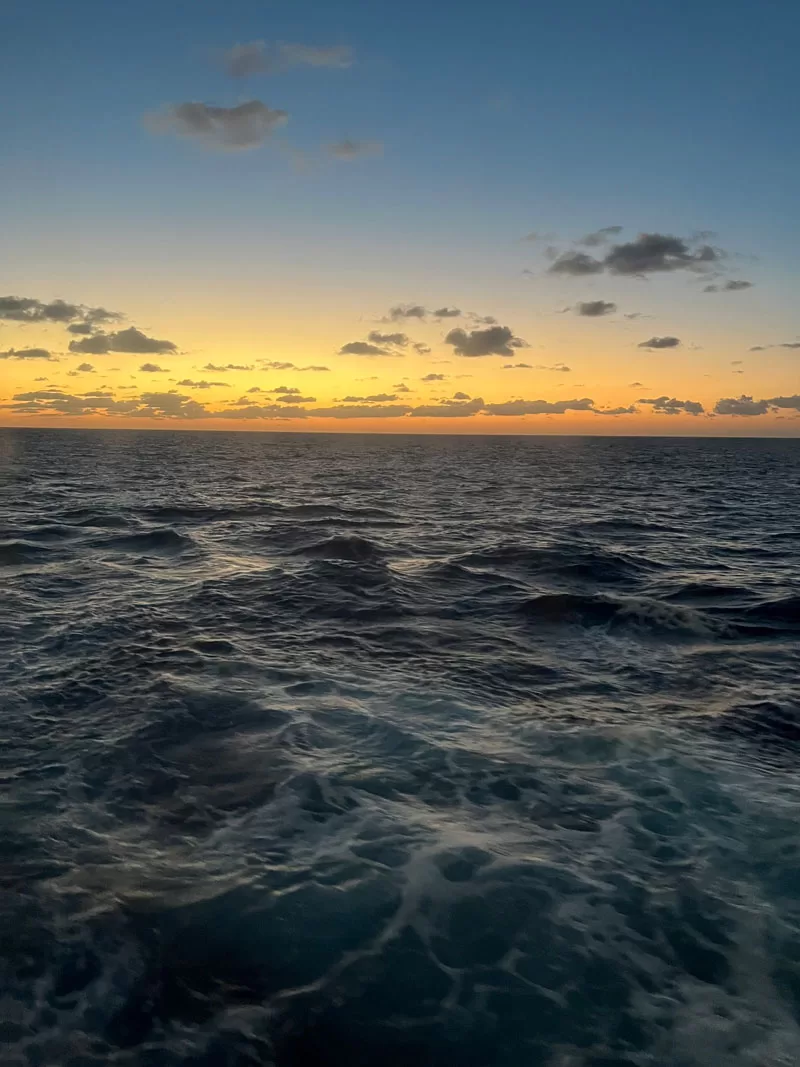 Ocean wake behind a cruise ship at sunset with blue water and a golden sky on the horizon.