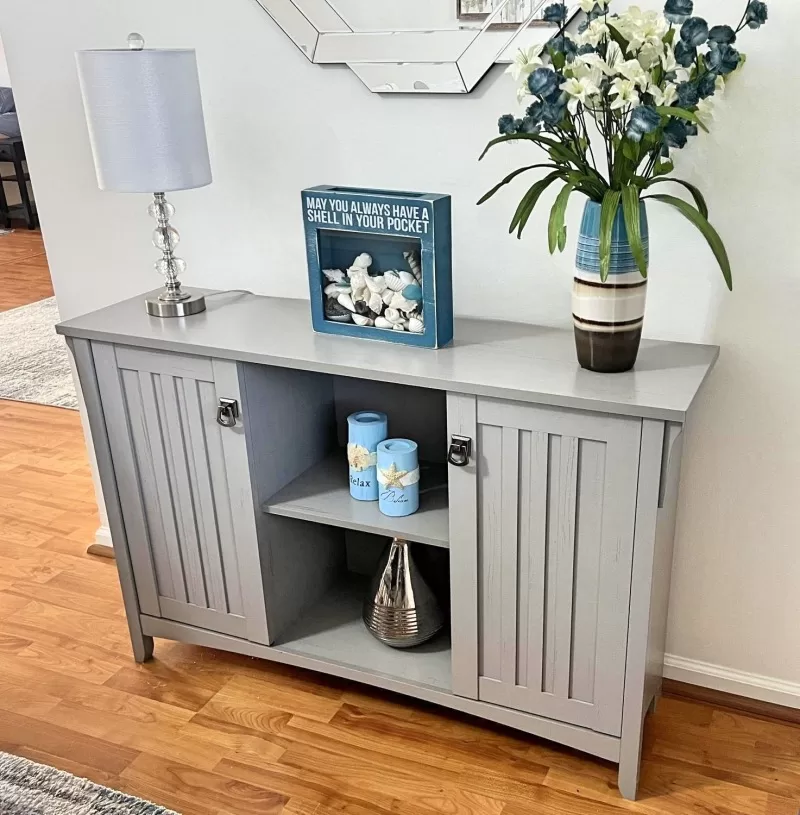 Grey buffet table styled with a crystal lamp, shell decor, blue candles, flowers, and a decorative mirror above.