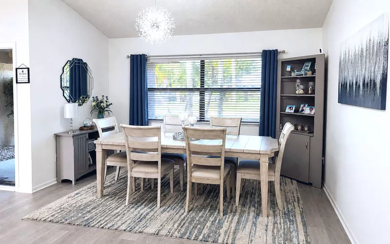 Updated dining room with grey table, blue curtains, crystal dandelion chandelier, buffet table, mirror, and painted corner cabinet.