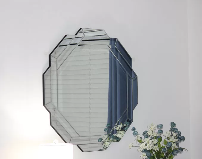 Decorative hexagon mirror on a white wall reflecting blue curtains, with flowers and a lamp below.