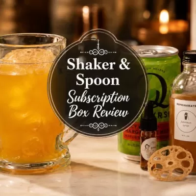 Orange craft cocktail with Shaker & Spoon mixer, syrup bottle, bitters, and dried garnish on a warm home bar counter