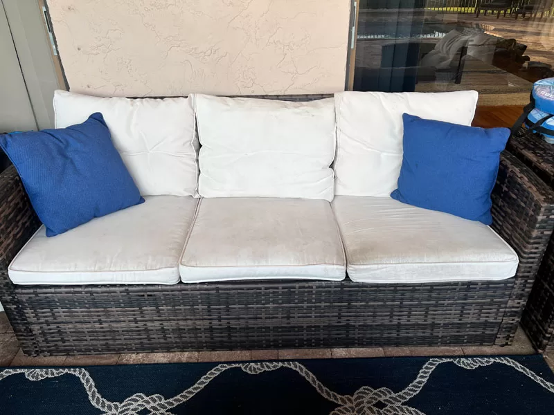 Old cream patio cushions on a dark wicker outdoor sofa with blue throw pillows before replacement covers