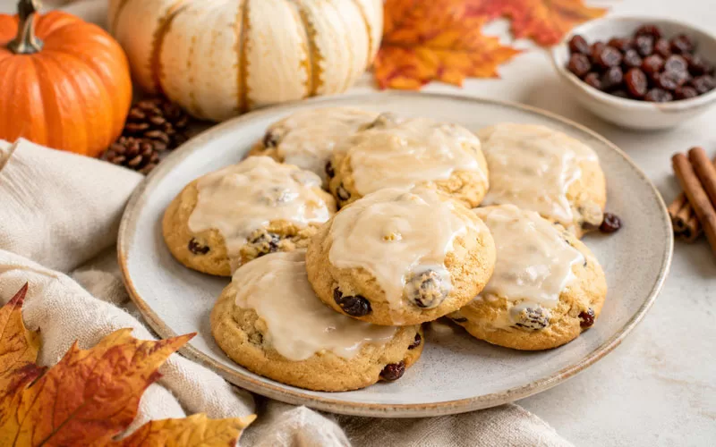 Glazed pumpkin raisin cookies on a plate with fall pumpkins, cinnamon sticks, raisins, and autumn leaves.