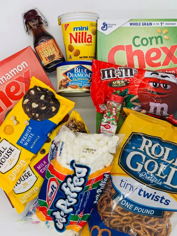 Flat lay of ingredients for Festive Sweet Maple Christmas Chex Mix including Corn Chex cereal, pretzels, marshmallows, vanilla wafers, almonds, maple syrup, chocolate chips, M&M’s, and holiday sprinkles.