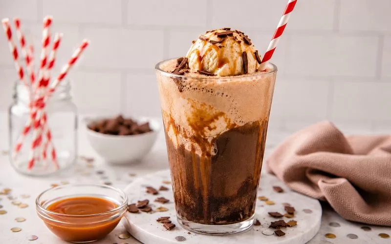 Salted caramel brownie float topped with vanilla ice cream, caramel drizzle, and chocolate shavings in a tall glass with striped straw.