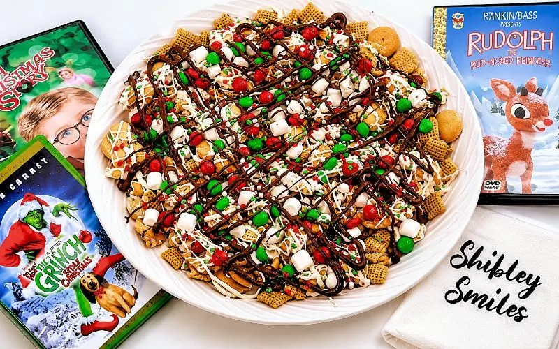 Holiday Sweet Maple Chex Mix recipe served on a white platter with chocolate drizzle, marshmallows, festive candies, and classic Christmas movie DVDs nearby.