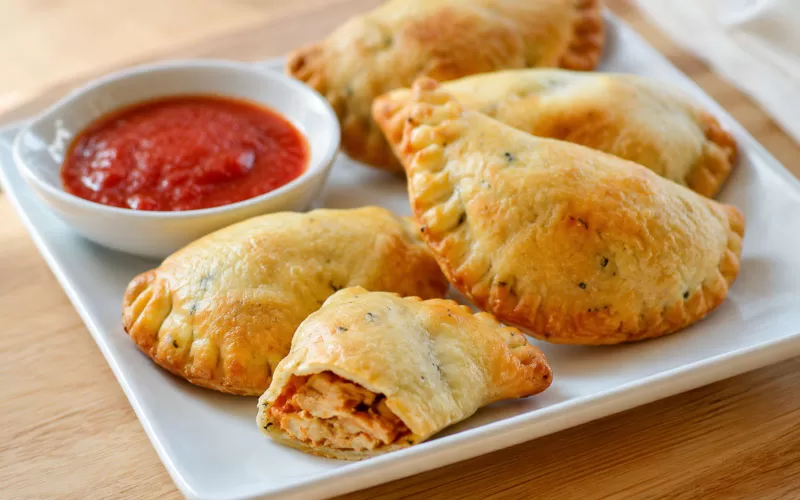 Golden chicken empanadas served on a white plate with red dipping sauce and one empanada opened to show the chicken filling.