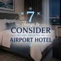 Modern hotel room with a made bed, rolling suitcase, charging phone, and airport runway lights outside the window.