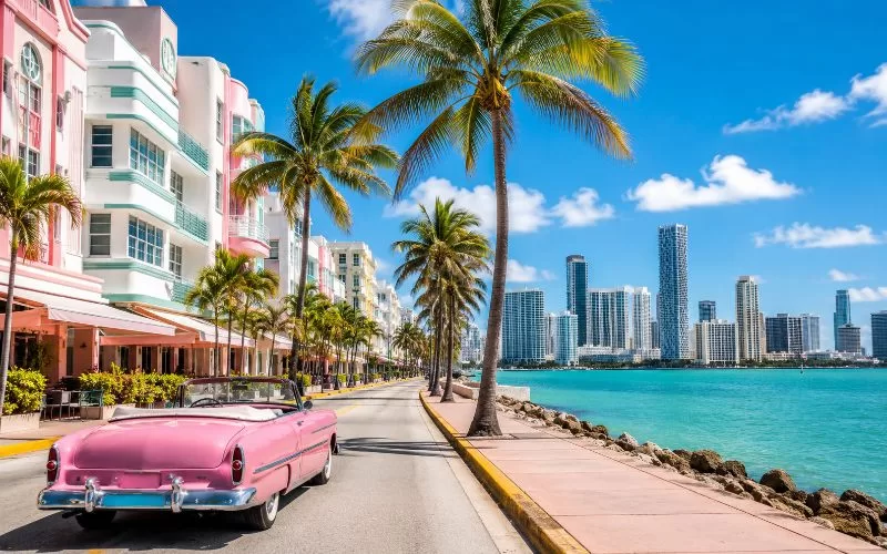 Scenic Miami drive with pastel Art Deco buildings, palm trees, turquoise water, skyline views, and a pink vintage convertible.