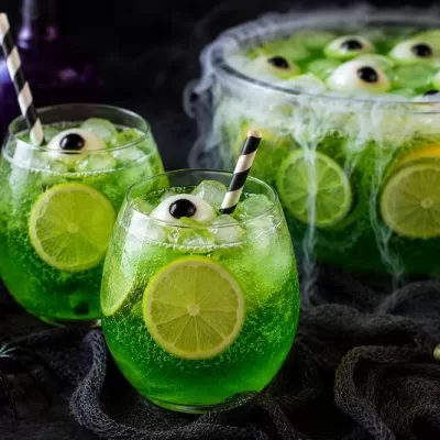 Two spooky green Halloween punch drinks with striped straws and candy eyeballs served beside a bubbling witches brew punch bowl with skull and spider decorations.