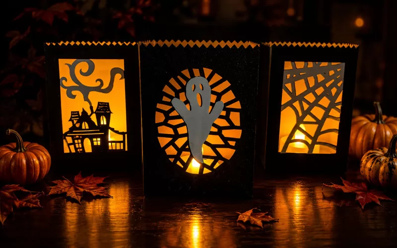 Glowing Halloween paper luminaries with haunted house, ghost, and spiderweb designs displayed with pumpkins and fall leaves on a dark tabletop.