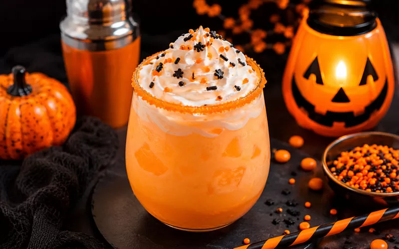Creamy orange Halloween Screamsicle cocktail topped with whipped cream and black and orange sprinkles surrounded by pumpkins and spooky Halloween decor.