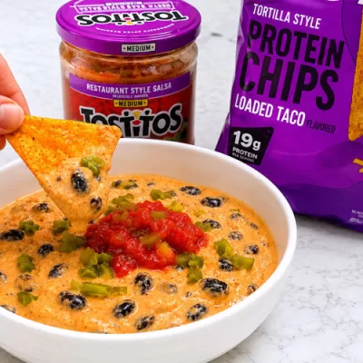Hand dipping a Quest loaded taco protein chip into a creamy cottage cheese queso dip topped with black beans, salsa, and diced jalapeños.