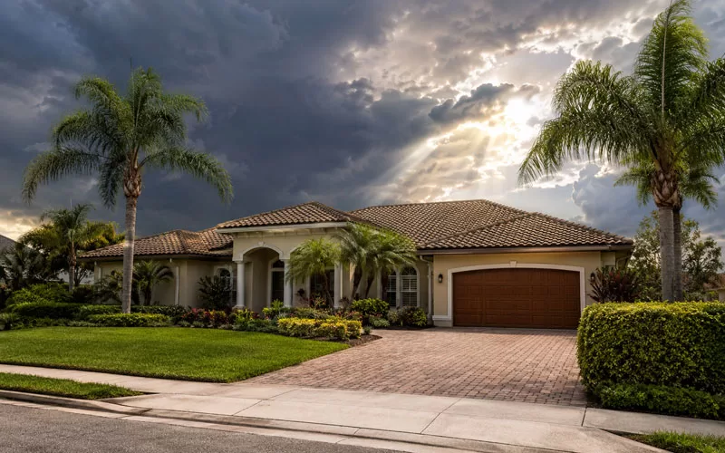 Florida home exterior with palm trees, a tiled roof, and dramatic storm clouds with sunlight breaking through.