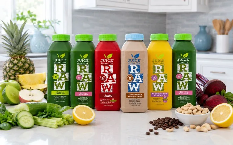 Juice From the Raw bottles arranged with pineapple, apples, cucumber, celery, lemon, beets, cashews, and coffee beans in a bright kitchen.